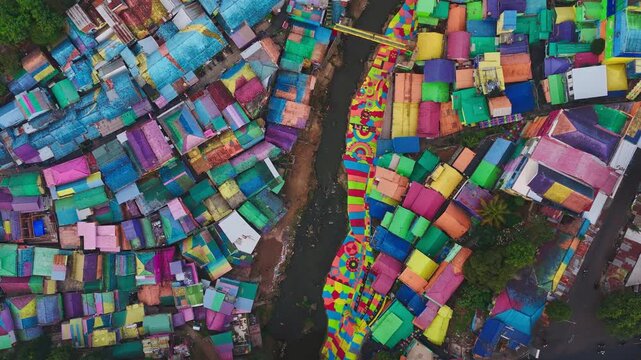 Colorful rooftops dominate this vibrant slum settlement. Winding paths cut through densely packed structures. Muddy river runs alongside the crowded dwellings. Buildings show varied hues