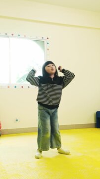 A young girl in a striped sweater dances on a yellow mat in a bright room near a window.