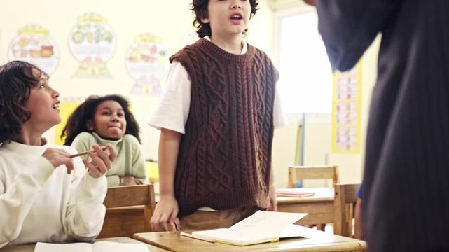 Children in a classroom listen as a student stands to speak while a teacher holds a laptop at the front of the room.
