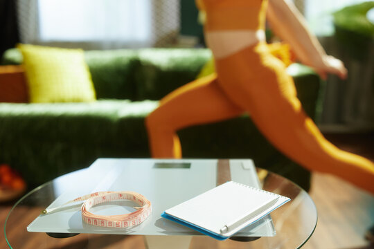 Focus on a digital scale, measuring tape, and notebook for progress tracking, with a woman in orange activewear performing lunges in a bright, sunlit living room.