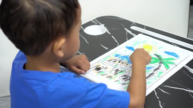 A small Asian boy is focusing on coloring a picture of a mountain landscape with crayons, to test the child's creativity, imagination and development.