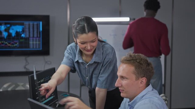 Female developer points to real-time performance metrics and AI insights on monitor while colleague reviews monitoring dashboard for technical analysis and optimization.