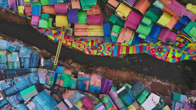 Colorful rooftops dominate this vibrant slum settlement. Winding paths cut through densely packed structures. Muddy river runs alongside the crowded dwellings. Buildings show varied hues