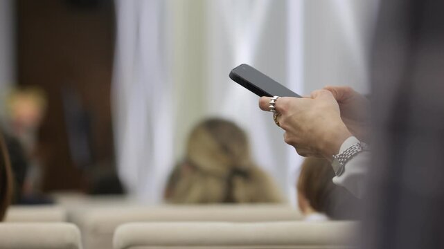 Person holds smartphone at event, ignoring speaker. Focus on hand and device. Blurred audience sits in rows behind. Shows modern distraction or multitasking in meetings. Useful for business, tech