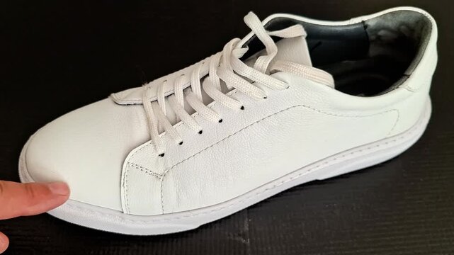 Finger points to scuff marks on white leather sneaker, highlighting wear and texture details, showing close up inspection of shoe surface for cleaning or product demo