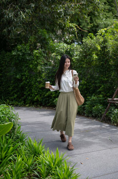 Young Asian woman smiling while looking at smartphone and holding coffee cup while walking in park.
