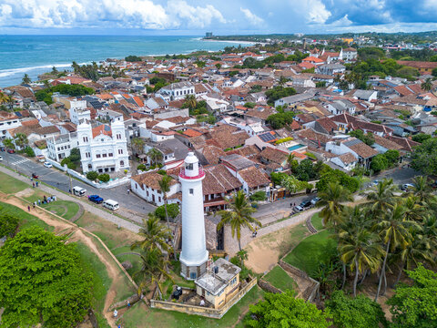 High-angle aerial view of the historic white Galle Lighthouse standing along the stone ramparts next to the Meeran Jumma Mosque and terracotta roofed buildings in Galle Fort, Sri Lanka.