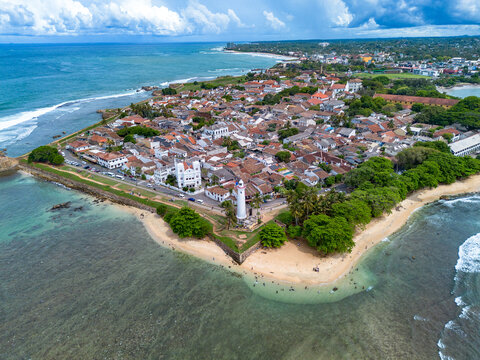 Beautiful aerial landscape view showing the tip of Galle Fort with its white lighthouse, sandy beach enclave, palm trees, and terracotta townscape framed by the Indian Ocean.