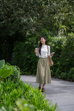 Young Asian woman carrying a coffee cup and phone while walking on footpath in the greenery park.