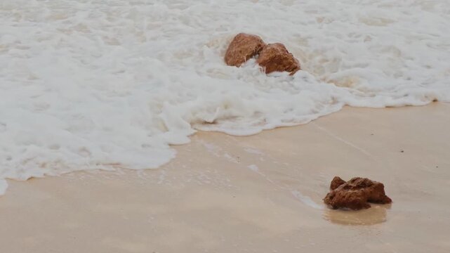 Relaxing video of ocean waves washing sandy shore with rocks and foam, perfect for nature and travel themes.