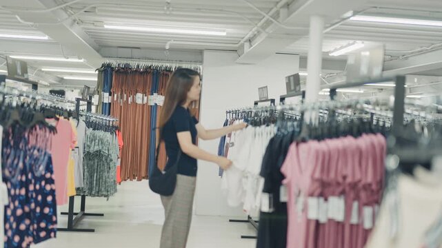 Young woman inspects a white blouse on a hanger while standing between clothing rails in a bright fashion store.