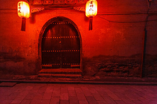 An old, arched doorway is illuminated by two hanging lanterns, casting a warm, red glow on the brick wall and stone steps, creating a sense of mystery and inviting exploration.