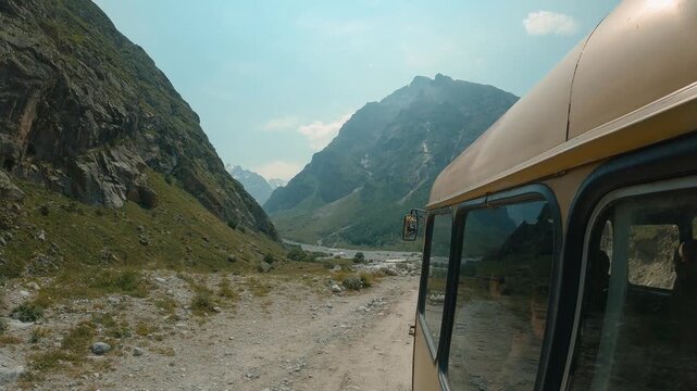 A dirt road winds through a green mountain valley with a rocky riverbed. Part of an old bus is visible on the right. In the distance, mountain peaks are visible in the soft afternoon light