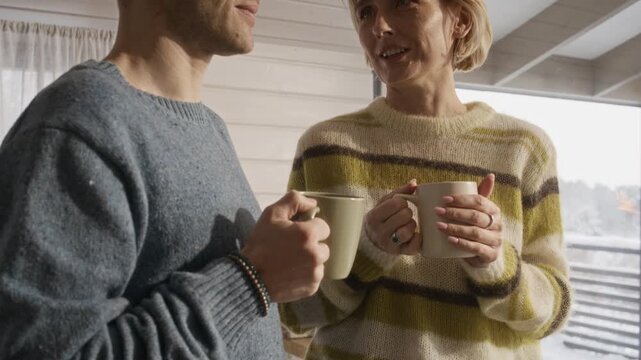 Middle aged Caucasian couple standing near window in wooden cabin, holding mugs and talking. Footage is perfect for real estate marketing, winter vacation promos, or family lifestyle campaigns