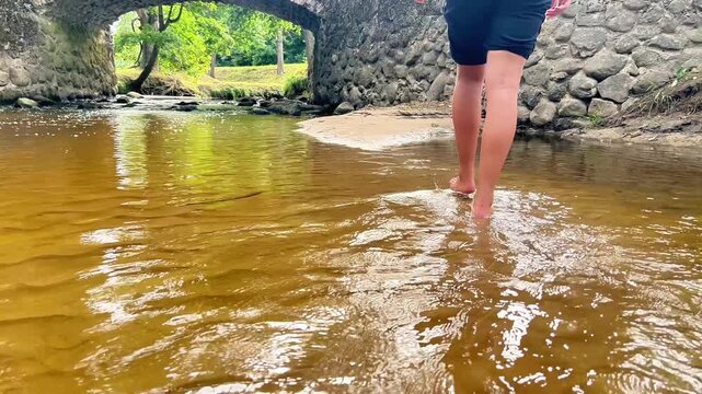 Low-angle shot of person walking barefoot through shallow river water near a historic stone bridge, ideal for eco-tourism, outdoor travel, and lifestyle concepts.
