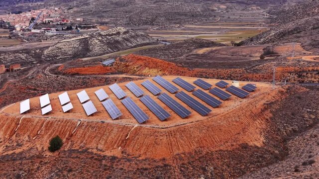 Solar farm installed on red rocky terrain surrounded by arid hills and distant farmland, rows of photovoltaic panels generating clean renewable energy in a rugged landscape