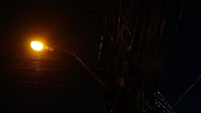 Dramatic low angle night view of tangled electric power cable wires on utility pole with glowing orange street lamp against dark blue sky