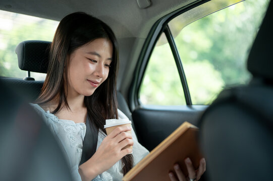 Young Asian woman holding coffee and reading a book while riding in the back seat of a taxi cab car.