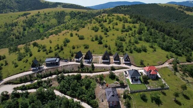 A drone passes a row of dark-roofed mountain chalets below open grassy hills and scattered trees in bright daylight in Zlatibor, Serbia on a sunny day.