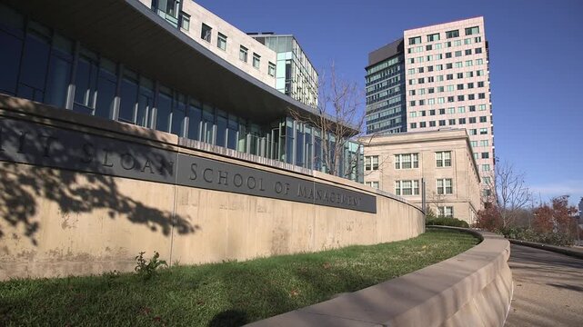 Exterior view of the MIT Sloan School of Management campus in Cambridge, Massachusetts, with modern academic buildings and a curved stone sign wall.
