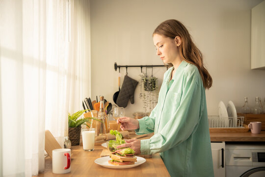 Young caucasian woman mixing fresh green salad at wooden counter near window. Side view of attractive female preparing healthy vegetable breakfast with sandwich and milk in kitchen.
