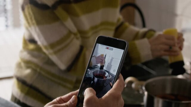 Middle aged Caucasian woman grinding pepper into pot while partner records her on smartphone in kitchen. Video suitable for food blogging, cooking apps, or lifestyle marketing campaigns