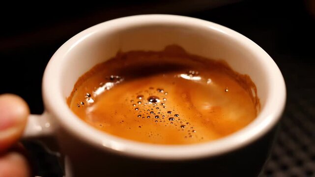 Closeup shot of freshly brewed espresso with rich brown crema in a small white cup, held by a hand. Dark background emphasizes the quality and texture of the coffee drink.