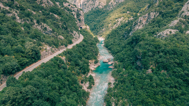 Aerial view discovering a scenic winding road through a lush green canyon with a vibrant turquoise river and a tunnel