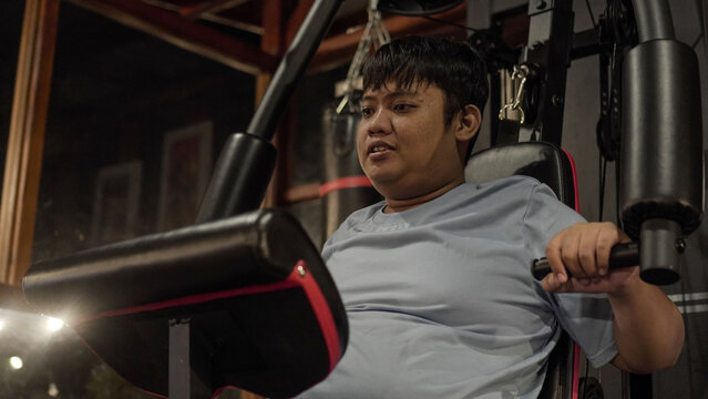 Exhausted Asian man struggling on a chest press machine in a night home gym.