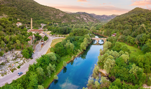 Rijeka Crnojevica river flowing past an old factory town and a stone arch bridge, surrounded by lush green mountains at sunset