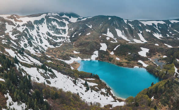 Alpine landscape with a turquoise Pesica lake surrounded by snow covered Bjelasica mountains, green trees, and cloudy skies in Montenegro