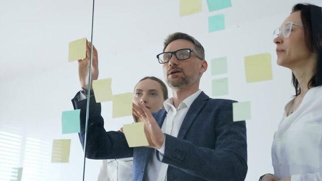 Business team collaboratively brainstorming new strategies and ideas on a glass whiteboard, creating a project plan with sticky notes and marker in a modern office meeting