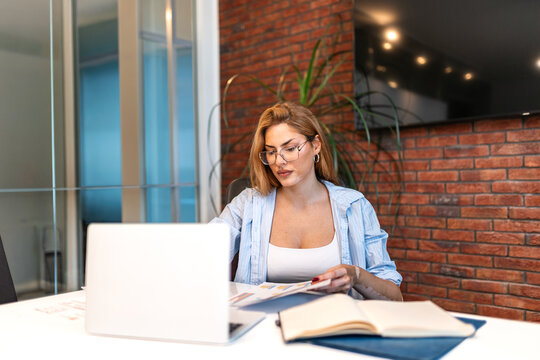 Focused businesswoman with vitiligo sitting at her office desk and reviewing business documents and graphs on paper, working diligently with a laptop and a whiteboard in the background.
