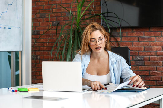 Young businesswoman with vitiligo working on laptop and analyzing financial reports in a modern office setup, surrounded by documents and a large brick wall background during daytime.