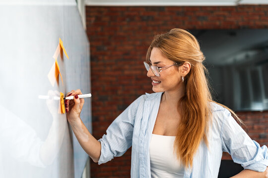 Smiling businesswoman with vitiligo writing colorful sticky notes attached to a clean white presentation board in a modern office, showcasing creative strategy and workflow planning.