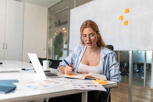 ocused young businesswoman with vitiligo sitting at office desk, wearing glasses and blue shirt while writing notes in notebook next to open laptop and financial charts.