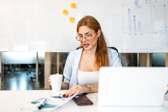 Businesswoman with vitiligo holding a white coffee cup while carefully checking financial documents at her bright office desk next to a laptop and smartphone during a busy workday.