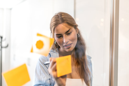 Focused young businesswoman with vitiligo writing ideas and notes on yellow sticky sticky notes attached to a glass wall in modern office during brainstorming session.