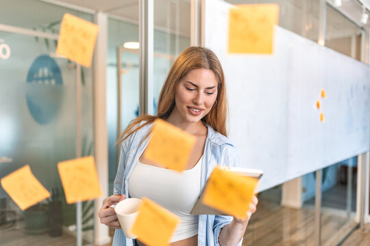 Smiling young businesswoman with vitiligo holding a digital tablet and coffee mug while reviewing project notes and sticky notes on a glass wall in a modern office space.