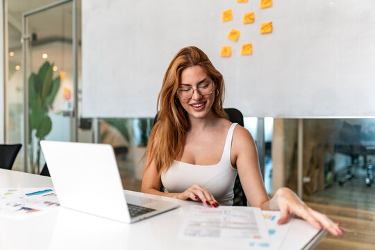 Smiling young businesswoman with vitiligo wearing glasses and white top, reviewing financial charts and documents at office desk. Corporate business administration and data analysis workflow concept.