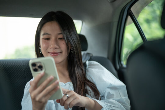 Young Asian woman smiling while checking smartphone in the back seat of a taxi cab car during a trip