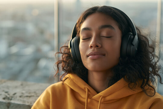 An intimate, eye-level close-up portrait of a young woman with curly hair, eyes closed, wearing headphones and enjoying music at sunset.
