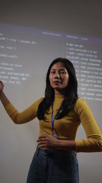 Professional woman developer presenting code on projection screen in tech training environment. confident expression, open hand gesture indicating active teaching or presentation.