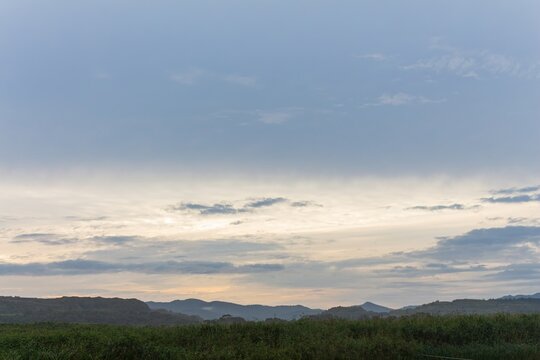 A vast, dramatic sky takes up most of the frame, hovering above a dark green foreground and a distant ridge of rolling hills, with soft golden light peeking through the cloud layers