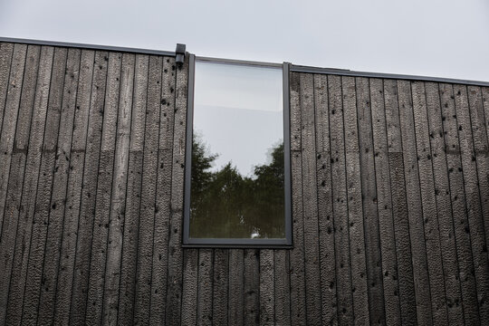 Low-angle view of a modern facade clad in black Shou Sugi Ban timber, featuring vertical charred boards, a tall reflective window, trees, and ample sky copy space