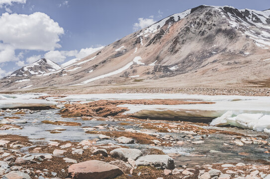 Landscape in the high Pamirs with rocky peaks and a river in a valley with melting snow. Panorama of rocky mountains on a sunny day and glaciers in a river valley