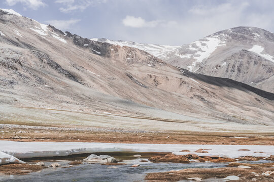 Landscape in the high Pamirs with rocky peaks and a river in a valley with melting snow. Panorama of rocky mountains on a sunny day and glaciers in a river valley