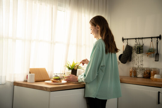 Wide angle view of young woman preparing healthy breakfast in bright sunlit modern kitchen. Female standing by kitchen counter with curtained window making sandwich and salad in the morning.