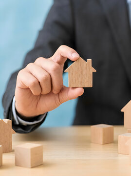 Businessman in a dark suit holding a small wooden house model on a wooden table with other wooden blocks representing real estate and construction