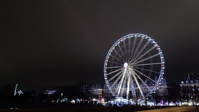 Night view of a giant illuminated ferris wheel lit up with blue and white lights against a dark night sky. Paris Christmas market carnival rides and historic city buildings in background.